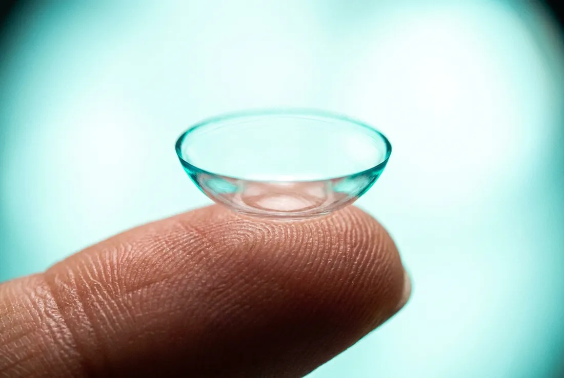 A single soft contact lens balanced on a fingertip, lit with a ring of teal light